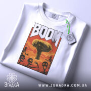 Футболка вибуховий арт BOOM білого кольору, складена на поверхні, фон світло-сірий, принт з ядерним вибухом та скелетами, на футболці є етикетка з зеленим шнурком.