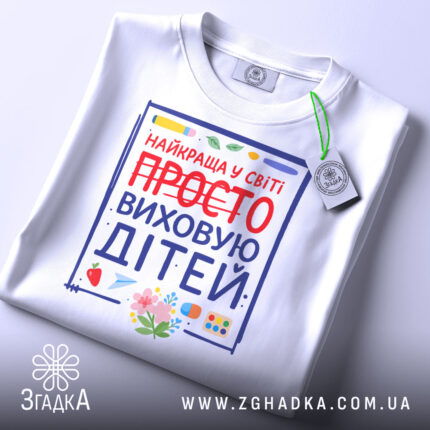 Футболка виховую дітей, біла, зображена зверху, фон світло-сірий, принт з написом 'НАЙКРАЩА У СВІТІ ПРОСТО ВИХОВУЮ ДІТЕЙ' з червоним закресленим словом 'ПРОСТО', деталі принта включають малюнки квітів та предметів для малювання.