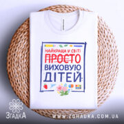 Футболка виховую дітей, біла, зображена зверху на плетеній кошику, фон світло-сірий, принт містить напис 'НАЙКРАЩА У СВІТІ ПРОСТО ВИХОВУЮ ДІТЕЙ' з червоним закресленим словом 'ПРОСТО', деталі принта включають малюнки квітів та предметів для малювання.