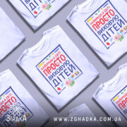 Футболка виховую дітей, біла, зображена з фронтального ракурсу, фон світло-сірий, принт містить яскраві кольори з написом 'НАЙКРАЩА У СВІТІ ПРОСТО ВИХОВУЮ ДІТЕЙ' та ілюстраціями фруктів і канцелярських предметів.