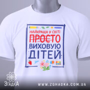 Футболка виховую дітей, біла, зображена зверху, фон світло-сірий, принт з написом 'НАЙКРАЩА У СВІТІ ПРОСТО ВИХОВУЮ ДІТЕЙ' з червоним закресленим словом 'ПРОСТО', деталі принта включають малюнки квітів та предметів для малювання.