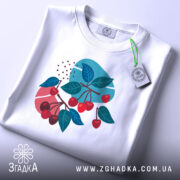 Футболка вишневі гілки, біла, складена на столі, фон світлий, принт з червоними вишнями та зеленим листям, з етикеткою на шнурку.