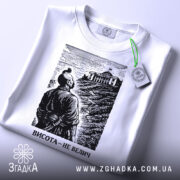 Футболка висота не велич. Біла футболка, складена на поверхні, з чорним принтом, що зображує чоловіка, який дивиться на будівлю. На футболці є етикетка з зеленим шнурком. Фон світлий, що підкреслює деталі принта.