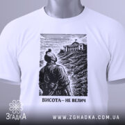 Футболка висота не велич. Біла футболка, складена на поверхні, з чорним принтом, що зображує чоловіка, який дивиться на будівлю. На футболці є етикетка з зеленим шнурком. Фон світлий, що підкреслює деталі принта.