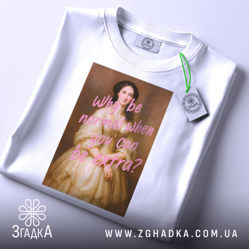 Футболка why be extra білого кольору, розкладена на поверхні, фон світло-сірий, принт з портретом жінки в бежевій сукні та текстом рожевого кольору, на комірці видна етикетка.