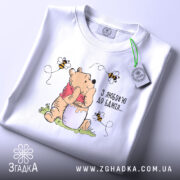 Футболка з любовʼю до бджіл, білого кольору, складена на поверхні, фон світлий, принт з ведмедиком, що тримає банку з медом, оточений бджолами, на футболці є етикетка.