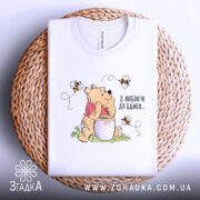 Футболка з любовʼю до бджіл, білого кольору, складена на плетеній основі, фон світлий, принт з ведмедиком, що тримає банку з медом, оточений бджолами.