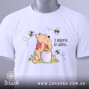 Футболка з любовʼю до бджіл, білого кольору, складена на поверхні, фон світлий, принт з ведмедиком, що тримає банку з медом, оточений бджолами, на футболці є етикетка.