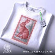 Футболка заєць лісовий, біла, зображена зверху, фон світло-сірий, принт з червоним зайцем на фоні лісу, з етикеткою зеленого кольору.