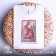 Футболка заєць лісовий, біла, розкладена на плетеній кошику, фон світло-сірий, принт з червоним зайцем на фоні лісу, видно деталі коміра.