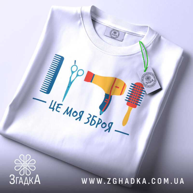 Футболка зброя перукаря, біла, зображена зверху, фон світло-сірий, принт містить гребінець, ножиці, фен та щітку, яскраві кольори, на футболці є етикетка.