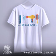 Футболка зброя перукаря, біла, підвішена на вішалці, фон світло-сірий, принт містить гребінець, ножиці, фен та щітку, яскраві кольори.