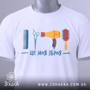 Футболка зброя перукаря, біла, зображена зверху, фон світло-сірий, принт містить гребінець, ножиці, фен та щітку, яскраві кольори, на футболці є етикетка.