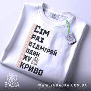 Футболка жарт про майстрів, біла, зображена зверху, фон світлий, принт містить текст "Сім раз відміряй один хукриво" та малюнок персонажа, лінійка розташована зліва, на футболці є етикетка з зеленим шнурком.