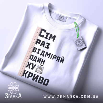 Футболка жарт про майстрів, біла, зображена зверху, фон світлий, принт містить текст "Сім раз відміряй один хукриво" та малюнок персонажа, лінійка розташована зліва, на футболці є етикетка з зеленим шнурком.