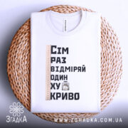 Футболка жарт про майстрів, біла, зображена зверху, фон світлий, принт містить текст "Сім раз відміряй один хукриво" та малюнок персонажа, лінійка розташована зліва, футболка лежить на плетеній основі.