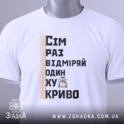 Футболка жарт про майстрів, біла, зображена зверху, фон світлий, принт містить текст "Сім раз відміряй один хукриво" та малюнок персонажа, лінійка розташована зліва, на футболці є етикетка з зеленим шнурком.