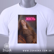 Футболка жесть класика, біла, складена на поверхні, фон світло-сірий, принт з дівчиною, яка дивиться вгору, з написом 'ЖЕСТЬ' у неоновому стилі, на футболці є етикетка з зеленим шнурком.