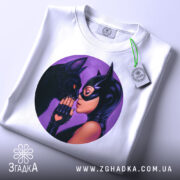 Футболка жінка котик, біла футболка, складена на поверхні, з круглим принтом, на якому жінка в масці торкається носа чорного кота, фон фіолетовий, на футболці є бірка з зеленим шнурком.