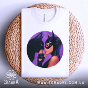 Футболка жінка котик, біла футболка, складена на плетеній основі, з круглим принтом, на якому жінка в масці ніжно торкається носа чорного кота, фон фіолетовий, ракурс зверху.