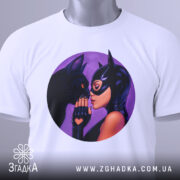 Футболка жінка котик, біла футболка, складена на поверхні, з круглим принтом, на якому жінка в масці торкається носа чорного кота, фон фіолетовий, на футболці є бірка з зеленим шнурком.