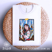 Футболка жіноча відвага, біла, складена на плетеній основі, фон світло-сірий, принт з дівчиною в українському вбранні, що тримає сокиру, на фоні дерев з червоними листям, з видимою етикеткою.