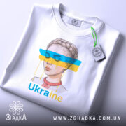 Футболка жіночий образ України, біла, зображена зверху, фон світло-сірий, принт з малюнком дівчини з косою, кольорові смуги синього і жовтого кольору, текст 'Ukraine' під малюнком, на футболці є етикетка з зеленим шнурком.