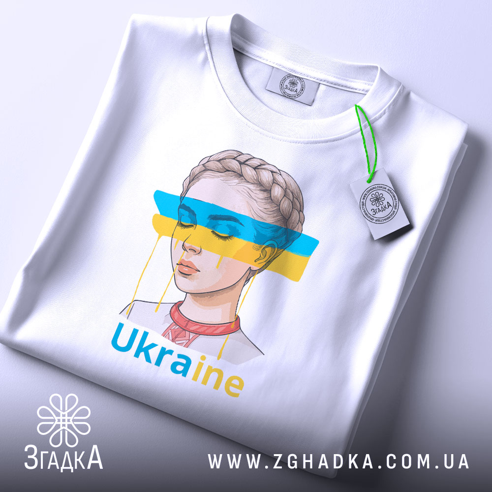 Футболка жіночий образ України, біла, зображена зверху, фон світло-сірий, принт з малюнком дівчини з косою, кольорові смуги синього і жовтого кольору, текст 'Ukraine' під малюнком, на футболці є етикетка з зеленим шнурком.
