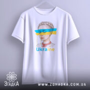 Футболка жіночий образ України, біла, зображена з бокового ракурсу, фон світло-сірий, принт з малюнком дівчини з косою, кольорові смуги синього і жовтого кольору, текст 'Ukraine' під малюнком, футболка висить на вішалці.