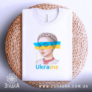 Футболка жіночий образ України, біла, зображена зверху на плетеній основі, фон світло-сірий, принт з малюнком дівчини з косою, кольорові смуги синього і жовтого кольору, текст 'Ukraine' під малюнком, видно етикетку на комірці.