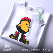 Футболка Жовта пташка, складена футболка з принтом жовтої пташки в червоних черевиках, фон світло-сірий, на футболці видно етикетку з логотипом.