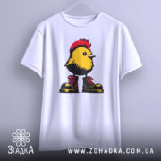 Футболка Жовта пташка, біла футболка на вішалці, з принтом жовтої пташки в червоних черевиках, фон світло-сірий, пташка має чорний гребінь і деталі на грудях.