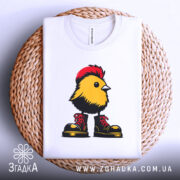 Футболка Жовта пташка, складена футболка на плетеній кошику, з принтом жовтої пташки в червоних черевиках, фон білий, видно етикетку з логотипом.