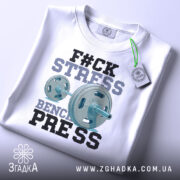 Футболка жим замість стресу, біла, складена на столі, фон світло-сірий, принт з написом 'F#CK STRESS' та зображенням гантелі, виконаний у блакитних та чорних кольорах, з етикеткою на зеленому шнурку.
