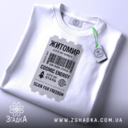 Футболка Житомир білого кольору, складена на поверхні, фон світло-сірий, принт у вигляді квитка з текстом 'Житомир', 'COSMIC ENERGY', 'SCAN FOR FREEDOM' у чорному кольорі, на комірці видніється етикетка.
