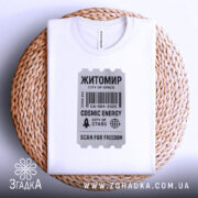 Футболка Житомир білого кольору, складена на плетеній кошику, фон світло-сірий, принт у вигляді квитка з текстом 'Житомир', 'COSMIC ENERGY', 'SCAN FOR FREEDOM' у чорному кольорі, видно етикетку на комірці.