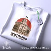 Футболка Житомир, біла, зображена зверху, на фоні світло-сірого градієнту, принт з зображенням будівлі з годинником та українським прапором на червоному фоні, текст 'Житомир' виконаний чорним шрифтом, на комірці видно етикетку.