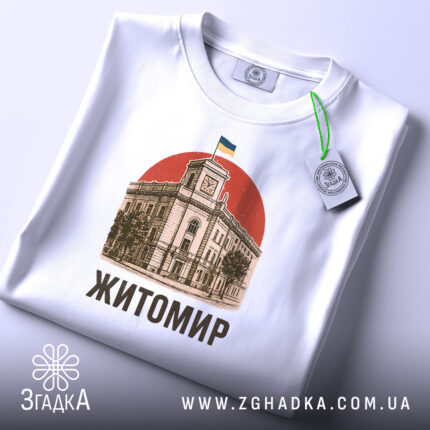 Футболка Житомир, біла, зображена зверху, на фоні світло-сірого градієнту, принт з зображенням будівлі з годинником та українським прапором на червоному фоні, текст 'Житомир' виконаний чорним шрифтом, на комірці видно етикетку.