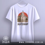 Футболка Житомир, біла, підвішена на вішалці, на фоні світло-сірого градієнту, принт з зображенням будівлі з годинником та українським прапором на червоному фоні, текст 'Житомир' виконаний чорним шрифтом.