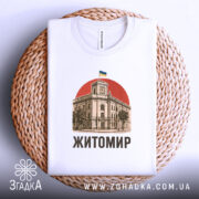 Футболка Житомир, біла, зображена зверху на плетеному кошику, на фоні світло-сірого градієнту, принт з зображенням будівлі з годинником та українським прапором на червоному фоні, текст 'Житомир' виконаний чорним шрифтом.