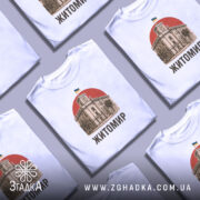 Футболка Житомир, біла, зображена з фронтального ракурсу, на фоні світло-сірого градієнту, принт з зображенням будівлі з годинником та українським прапором на червоному фоні, текст 'Житомир' виконаний чорним шрифтом.