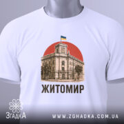 Футболка Житомир, біла, зображена зверху, на фоні світло-сірого градієнту, принт з зображенням будівлі з годинником та українським прапором на червоному фоні, текст 'Житомир' виконаний чорним шрифтом, на комірці видно етикетку.
