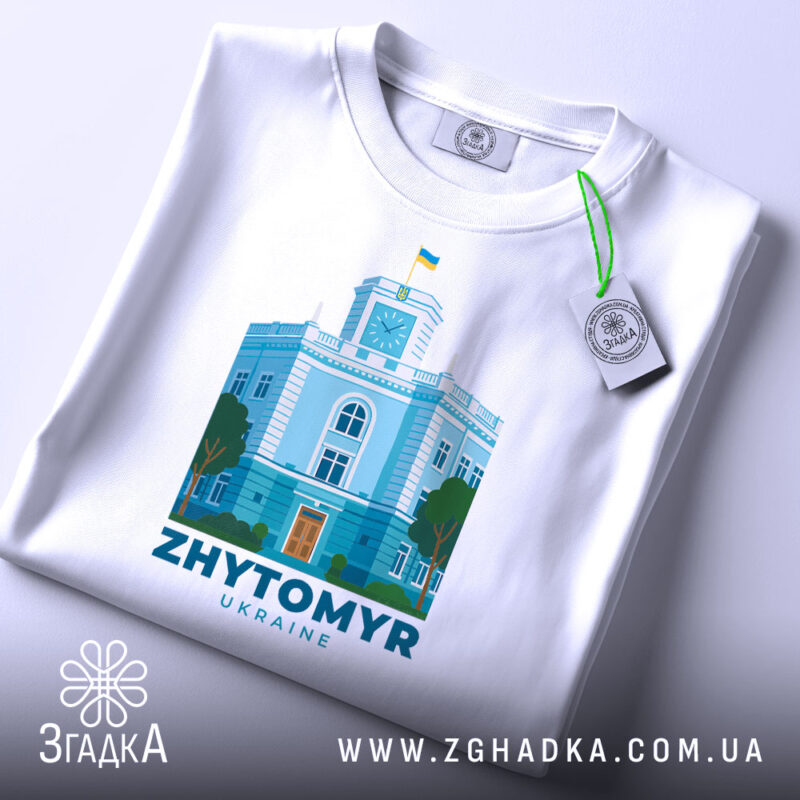 Футболка Житомир, біла, складена на поверхні, фон світлий, принт з зображенням будівлі з годинником та українським прапором, кольори принта: блакитний, зелений, помаранчевий, з етикеткою на шнурку.