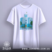 Футболка Житомир, біла, підвішена на вішалці, фон градієнтний сірий, принт з зображенням будівлі з годинником та українським прапором, кольори принта: блакитний, зелений, помаранчевий.
