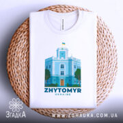 Футболка Житомир, біла, складена на кошику, фон світлий, принт з зображенням будівлі з годинником та українським прапором, кольори принта: блакитний, зелений, помаранчевий.