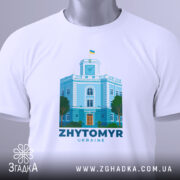 Футболка Житомир, біла, складена на поверхні, фон світлий, принт з зображенням будівлі з годинником та українським прапором, кольори принта: блакитний, зелений, помаранчевий, з етикеткою на шнурку.
