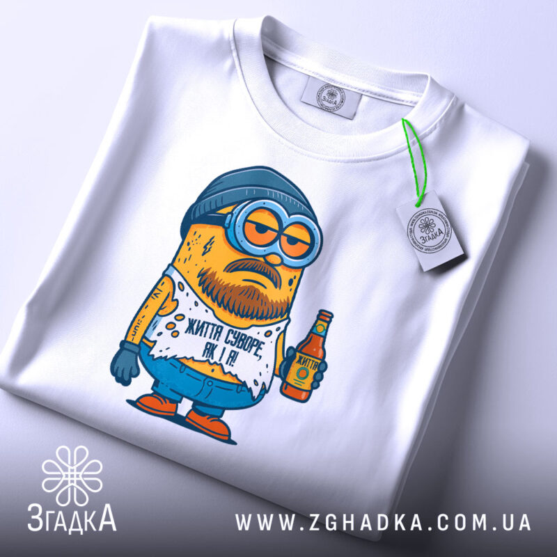 Футболка життя суворе, біла, складена на столі, фон світлий, принт з мультяшним персонажем у шапці та окулярах, тримає пляшку, текст на футболці: 'Життя суворе, як я!', з етикеткою на рукаві.