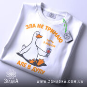 Футболка зла не тримаю, білого кольору, зображена на плоскій поверхні, фон світло-сірий, принт з качкою, що тримає келих з вином, текст оранжевого кольору, на футболці є етикетка з зеленим шнурком.