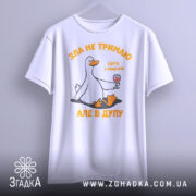 Футболка зла не тримаю, білого кольору, підвішена на вішалці, фон світло-сірий, принт з качкою, що тримає келих з вином, текст оранжевого кольору, вільний крій.