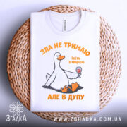 Футболка зла не тримаю, білого кольору, складена на плоскій поверхні з плетеного матеріалу, фон світлий, принт з качкою, що тримає келих з вином, текст оранжевого кольору, видима етикетка на горловині.